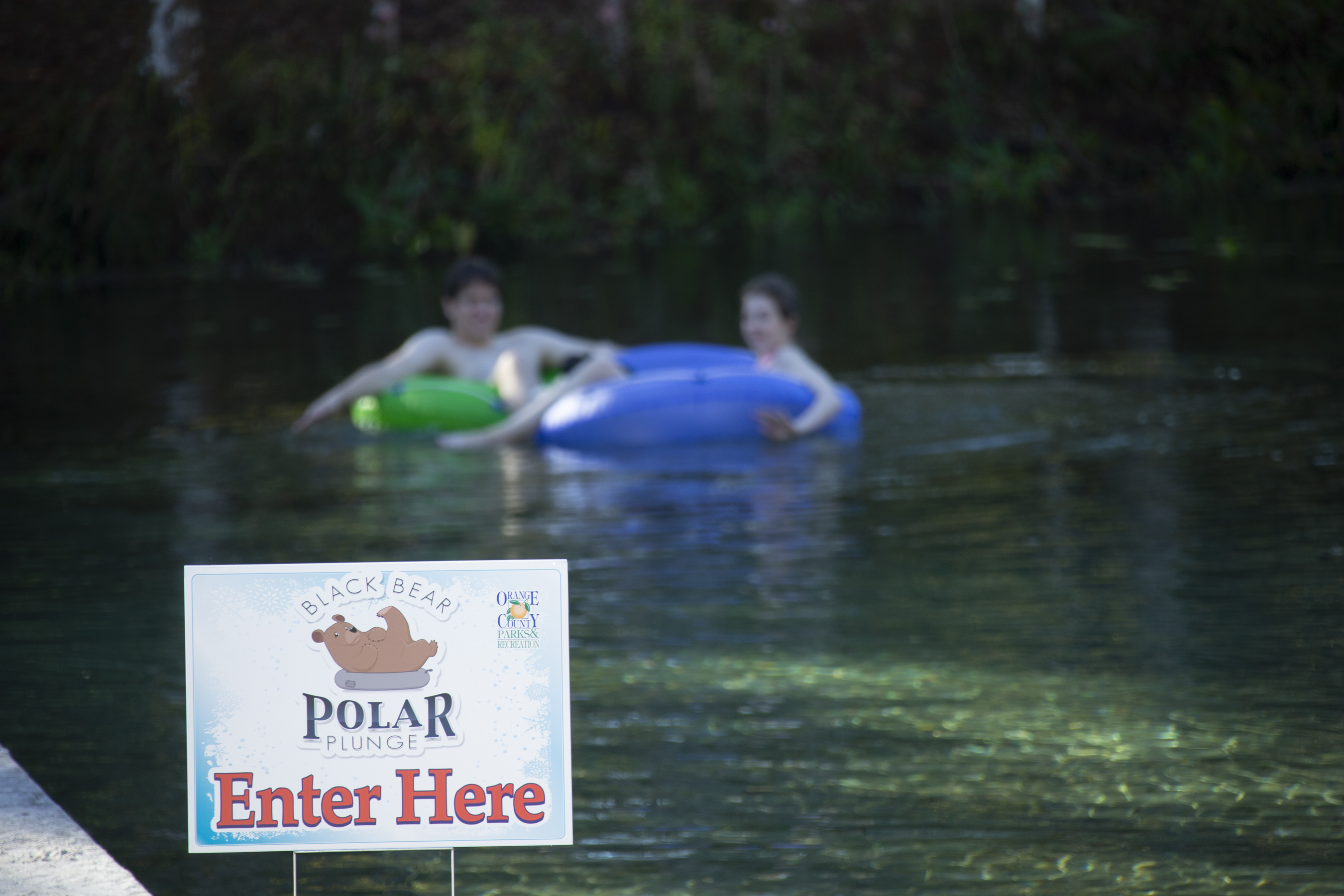 In the foreground there is a sign advertising the polar plunge event, while in the background there are two people on rubber donuts floating in the water.