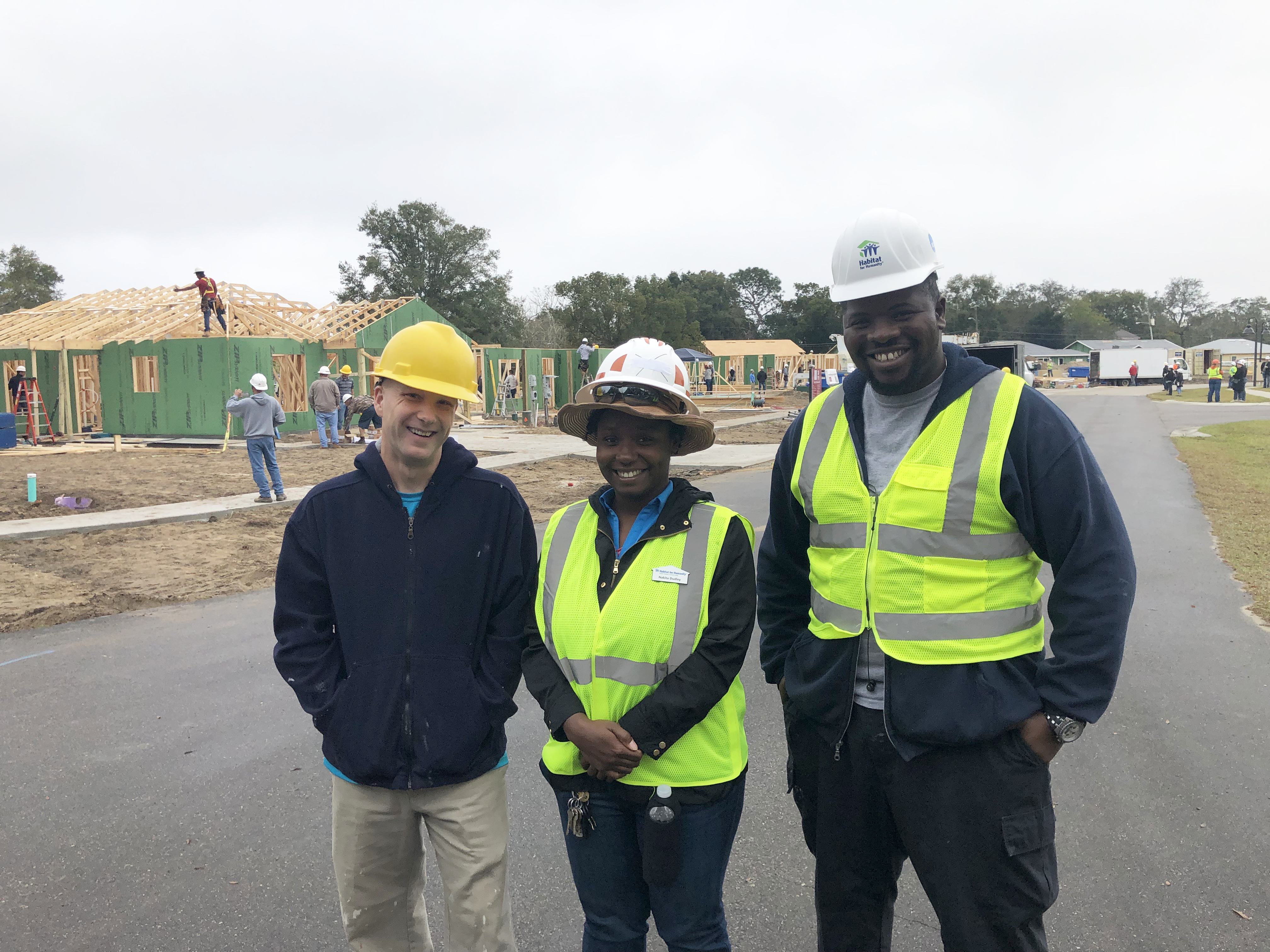 Three Habitat for Humanity employees wearing vests and hard hats pose for a photo. In the background, homes are actively being built.