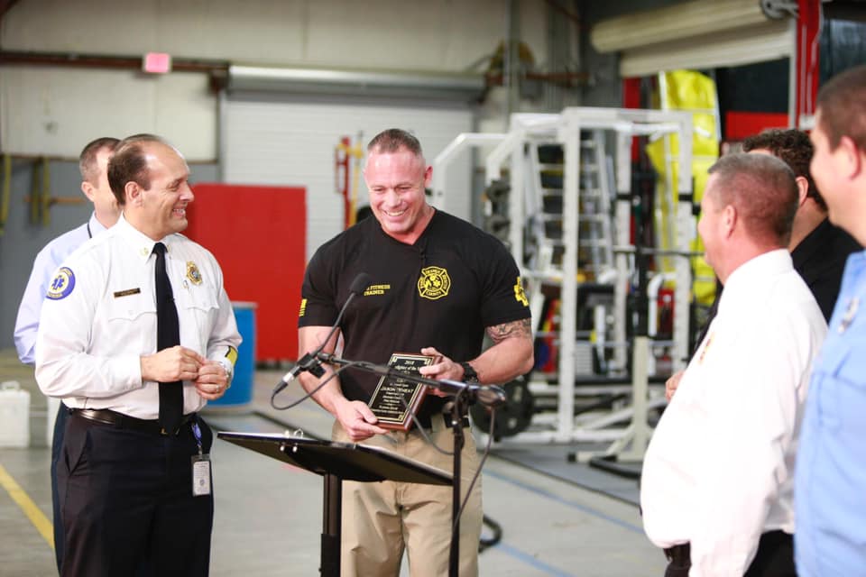 Inside of a Fire Station, firefighter Jason Wheat smiles while holding his Firefighter of the Year Award. Fire Chief Otto Drodz and other leaders smile as Jason walks towards the podium.