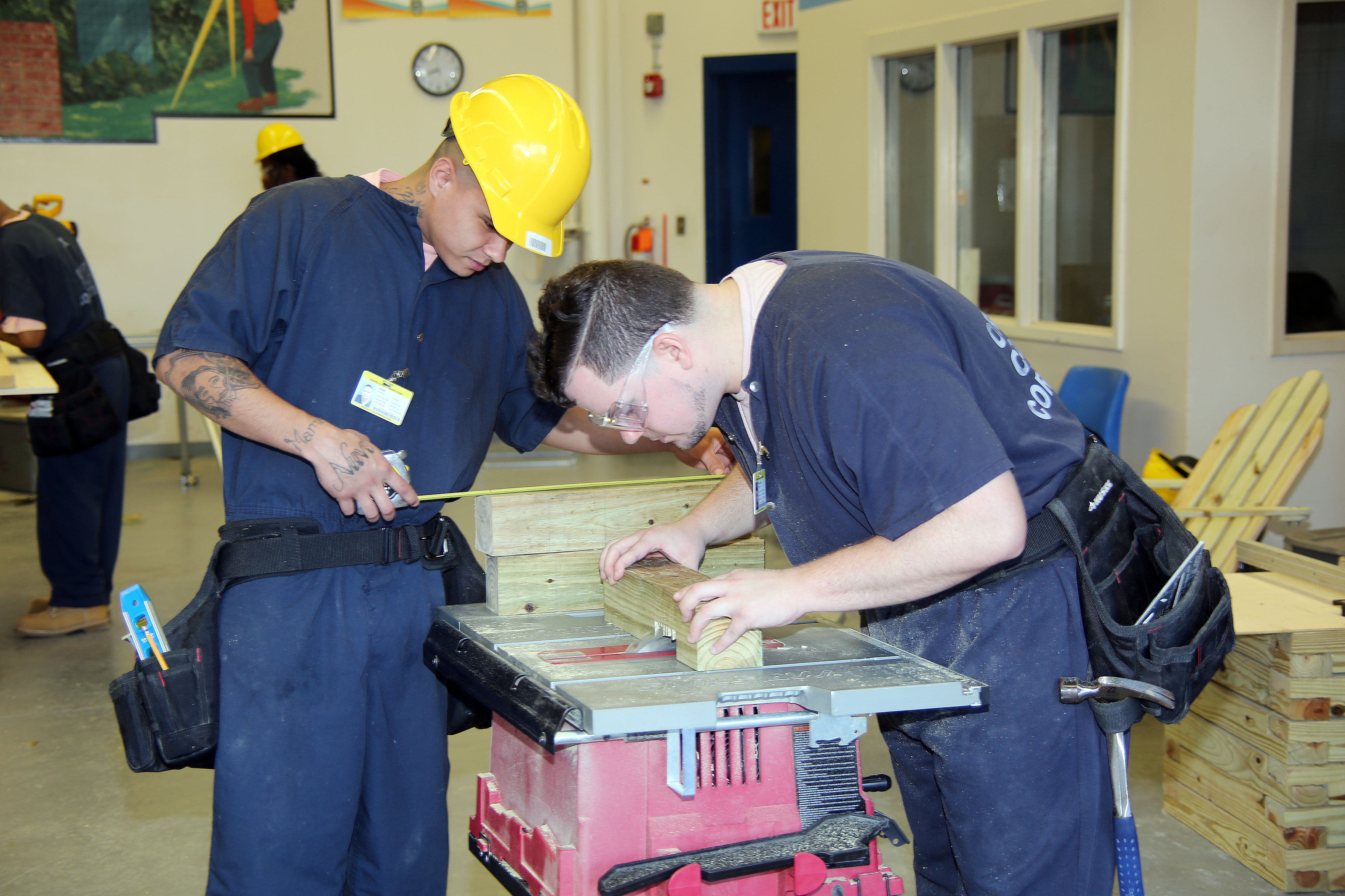 Two male inmates hunched over a woodworking table during their class at the corrections facility. They are intently focused on the project they are working on.