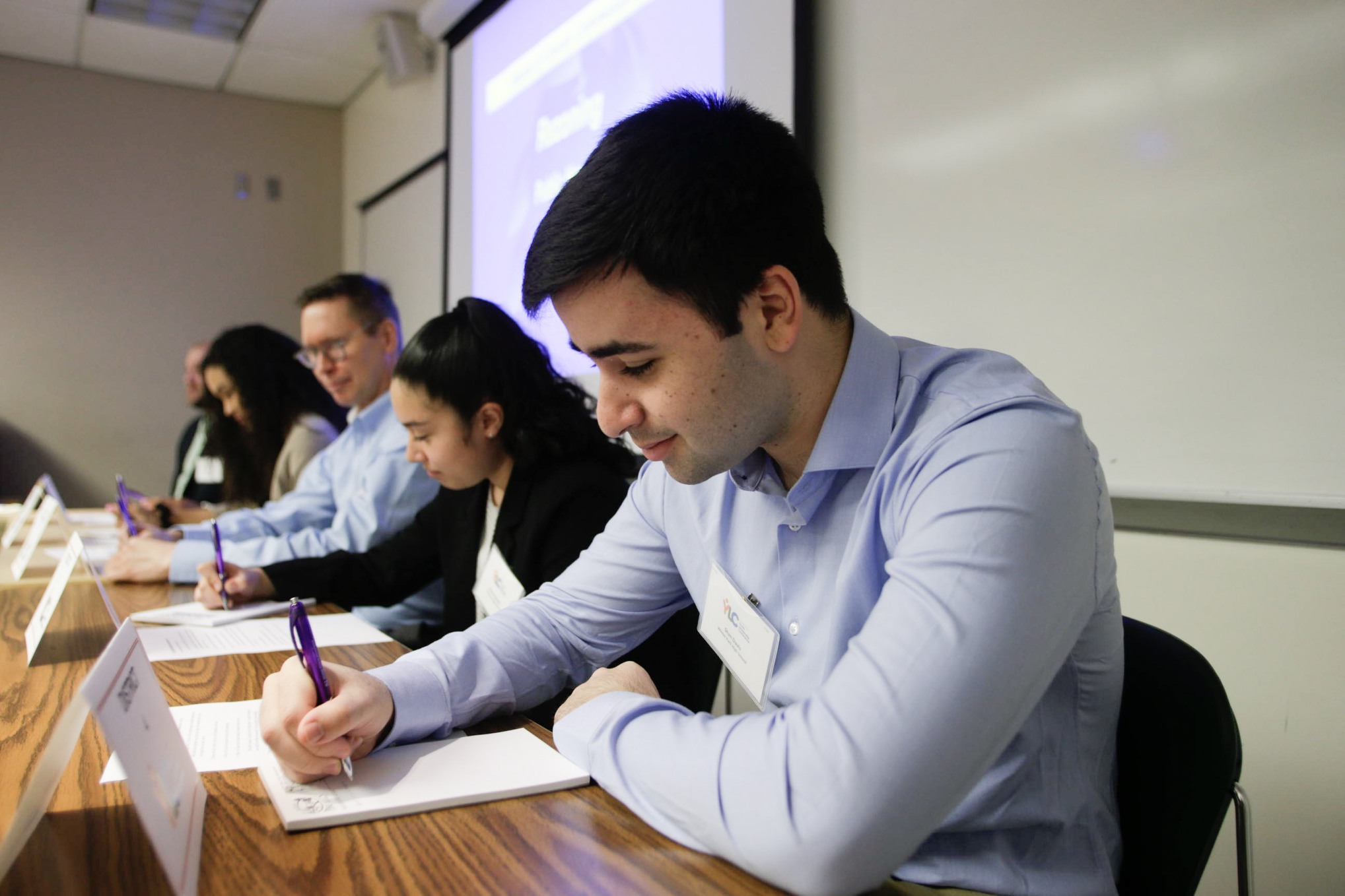 A close up shot of a student panelist focusing on a sheet of paper with pen in hand, as are the three other students and employees sitting alongside him.