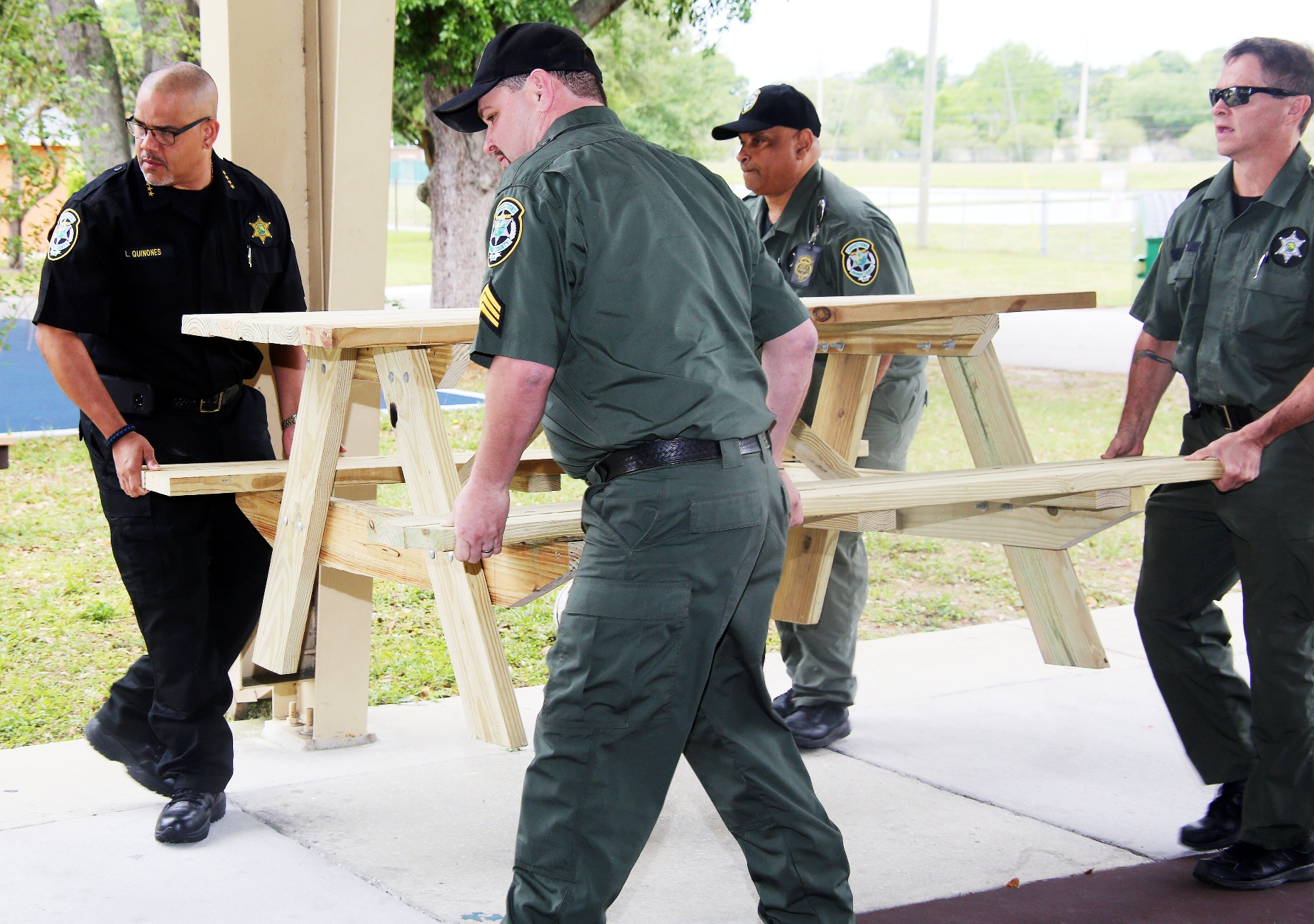Four corrections officers carrying a picnic table with benches. They are outside under a pavilion.