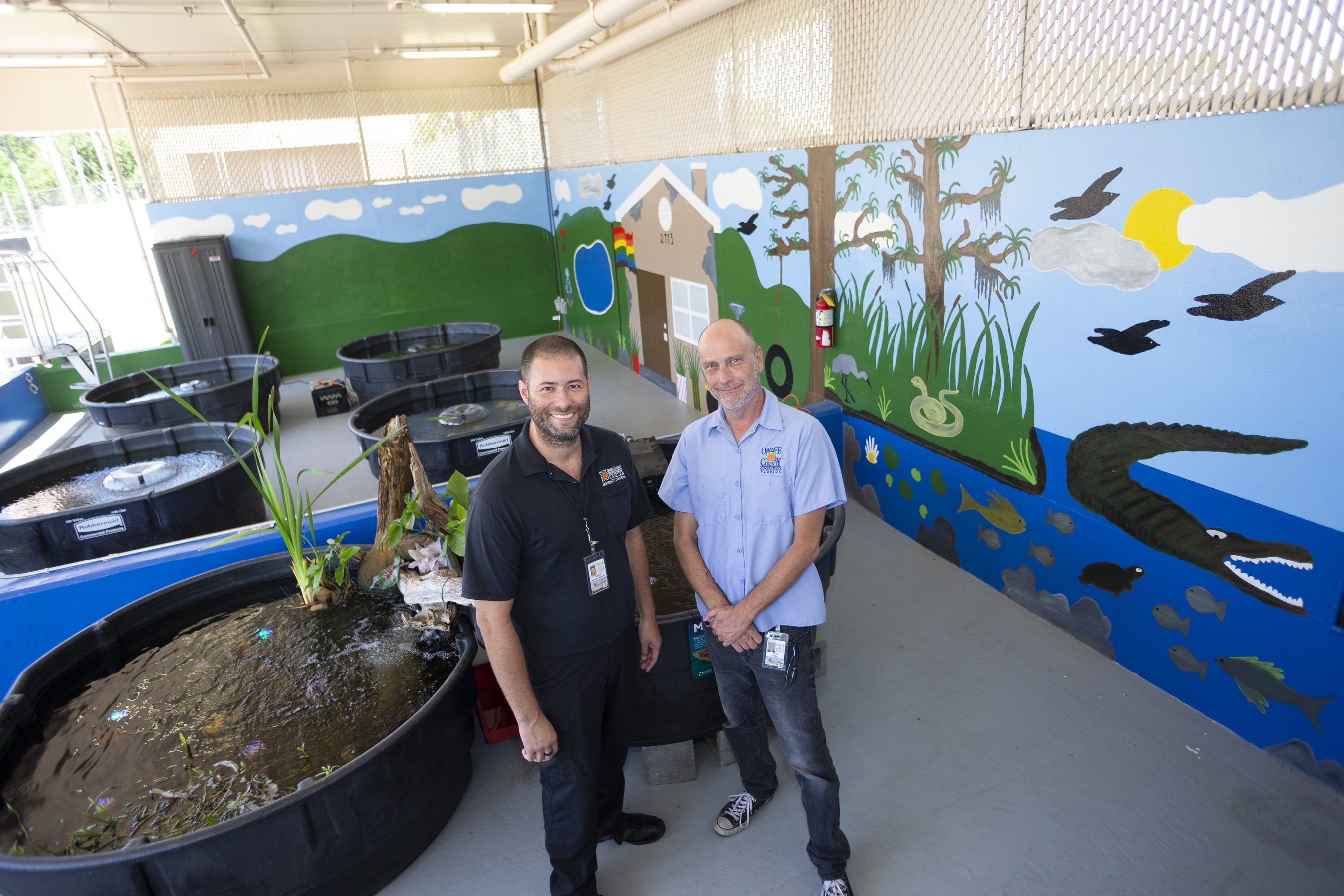 Two men inside a building, standing by six car-size containers full of water and fish. Behind them is a newly painted mural depicting a lake and wildlife.