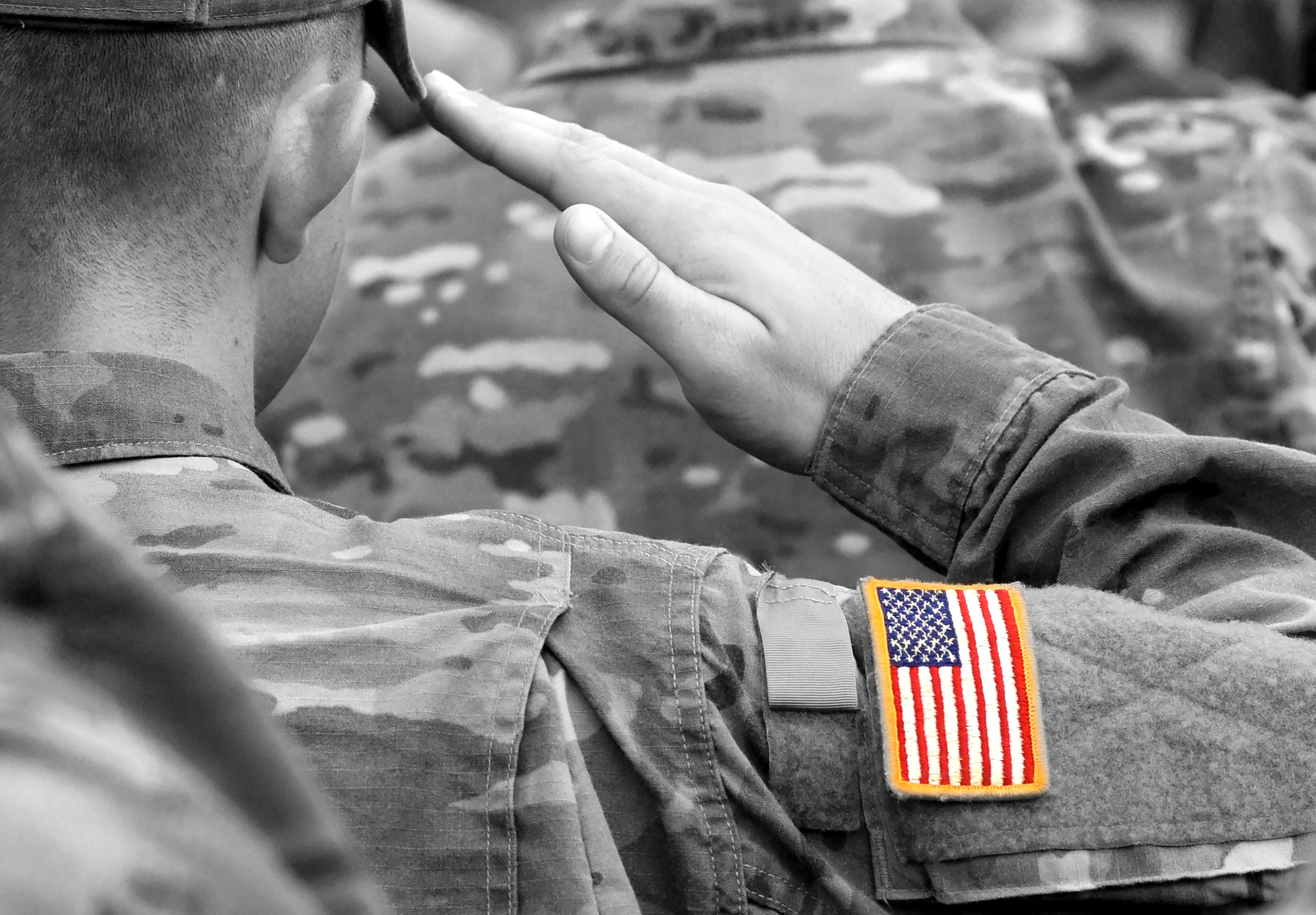 A close up photo of a man in a camouflage uniform saluting. On his sleeve the American flag is shown.