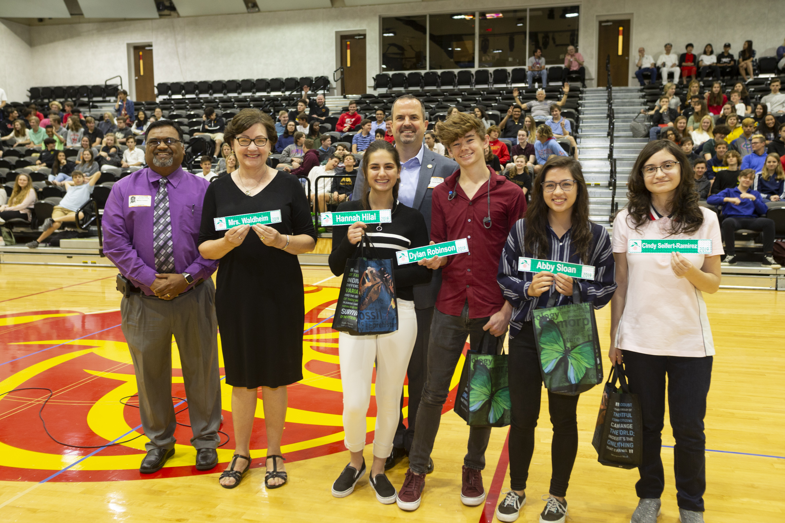 Three adults and four students standing in the middle of a school gym, smiling. The students are holding miniature road signs with their names on them.