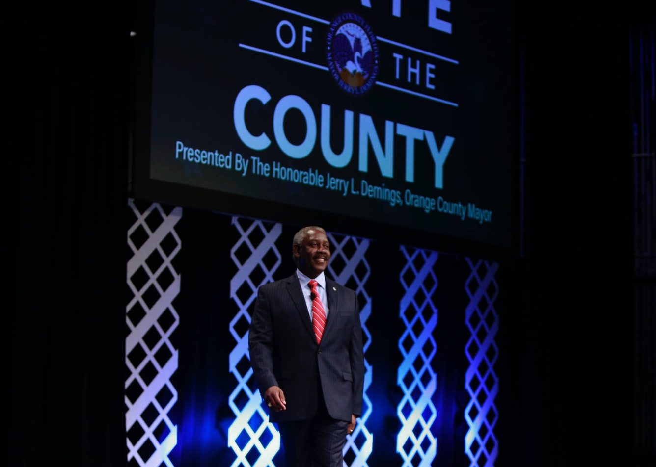 El Alcalde Demings parado en el escenario. Se puede leer "Estado del Condado" en el fondo.
