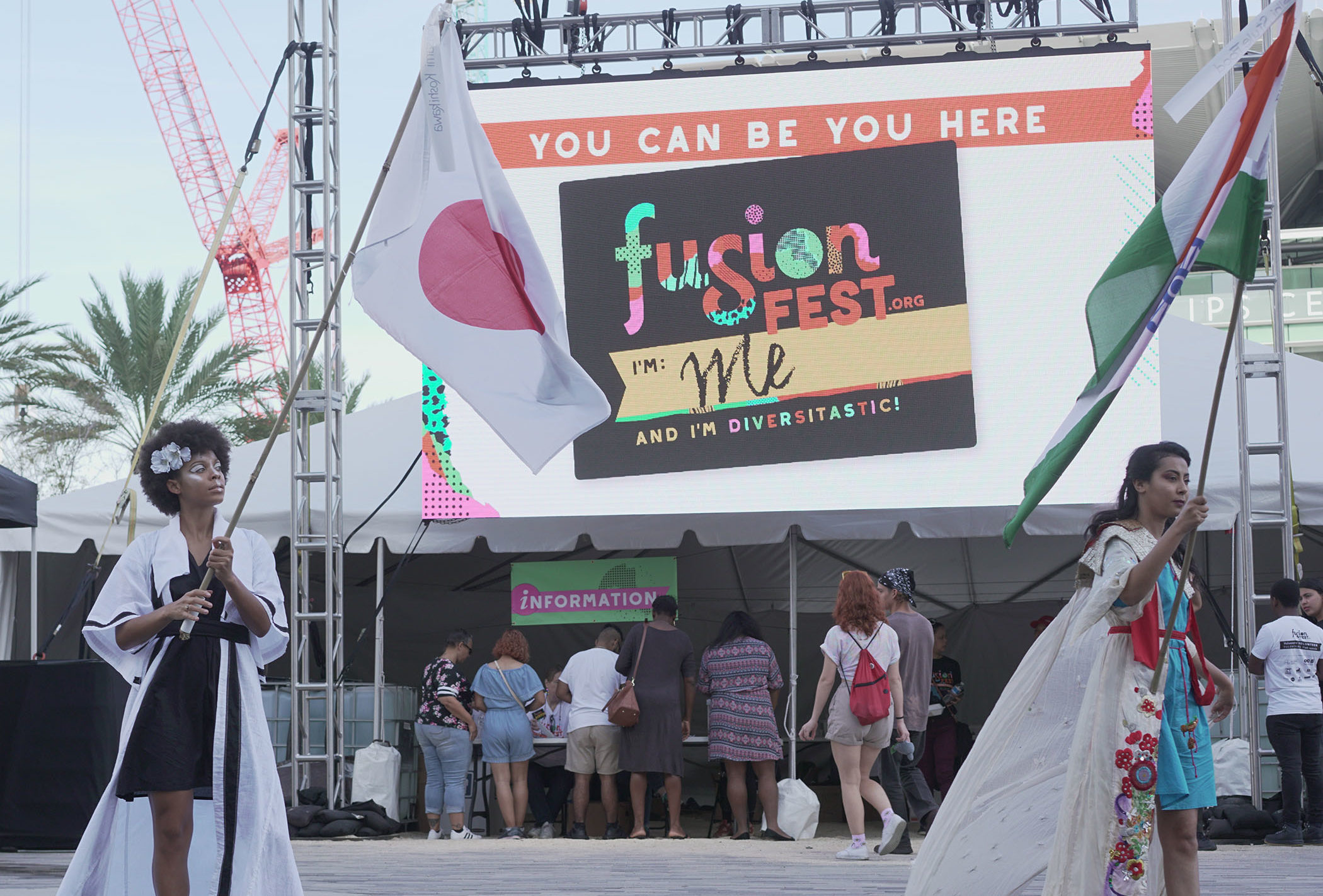Two women carrying flags of different nations on a pole. Behind them is a large screen that reads "You can be You here. Fusion Fest."