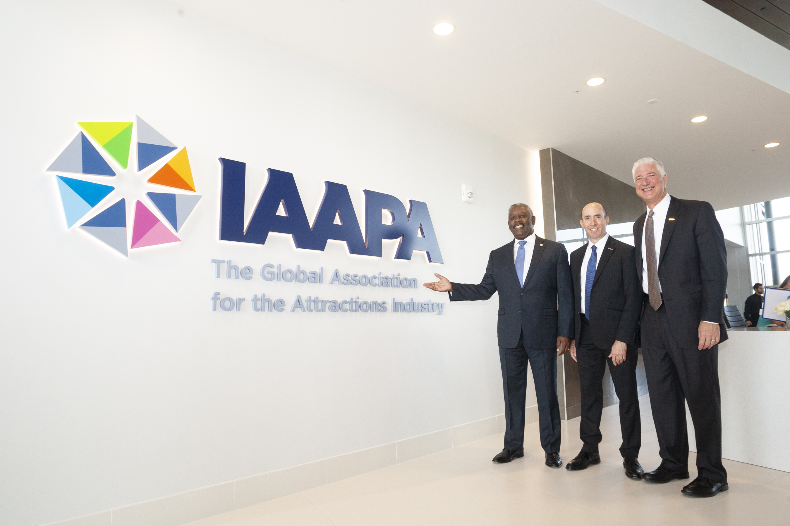 El Alcalde Demings y otros dos hombres parados junto a una pared con el logo de la IAAPA 