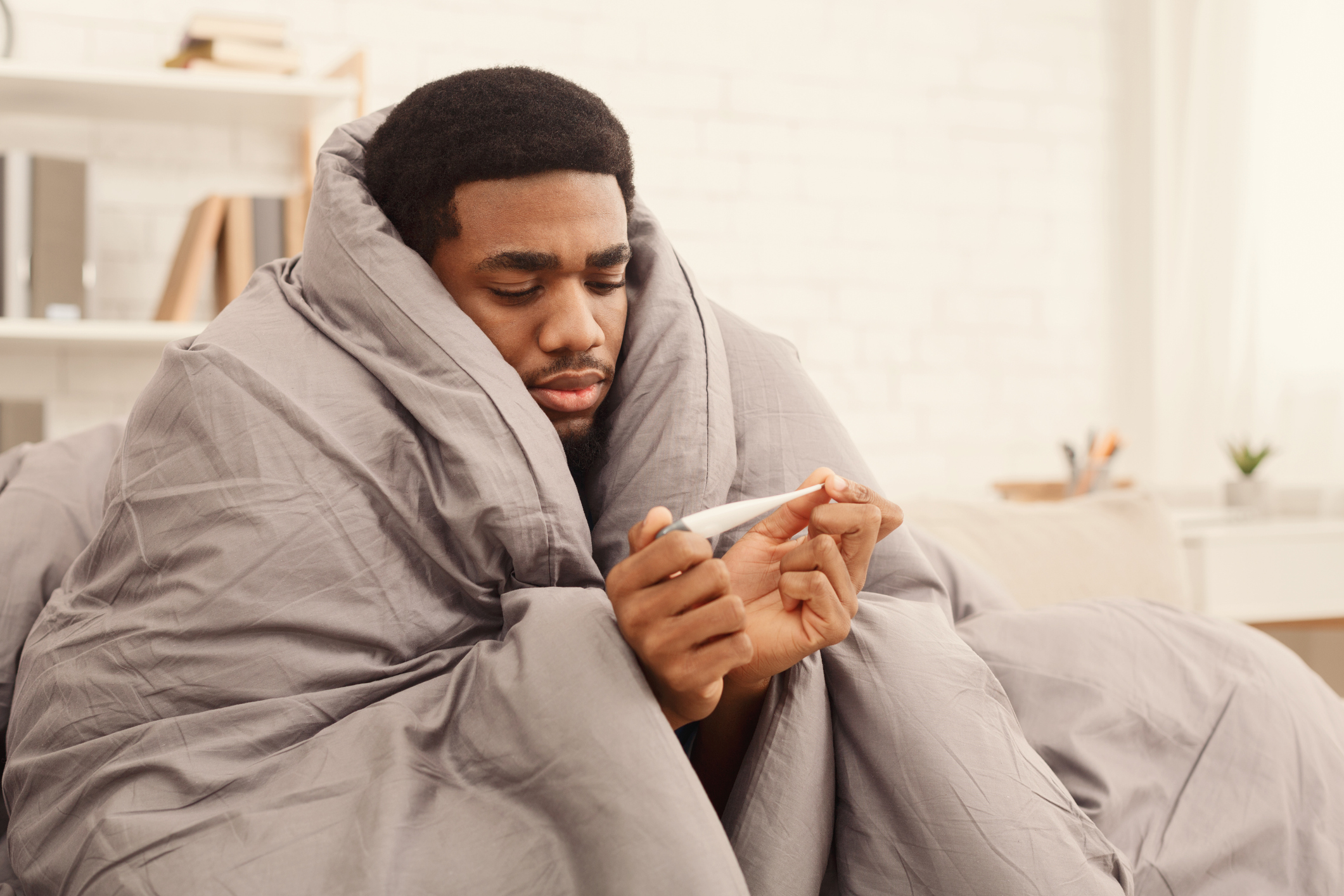 Ill man covered with blanket holding thermometer