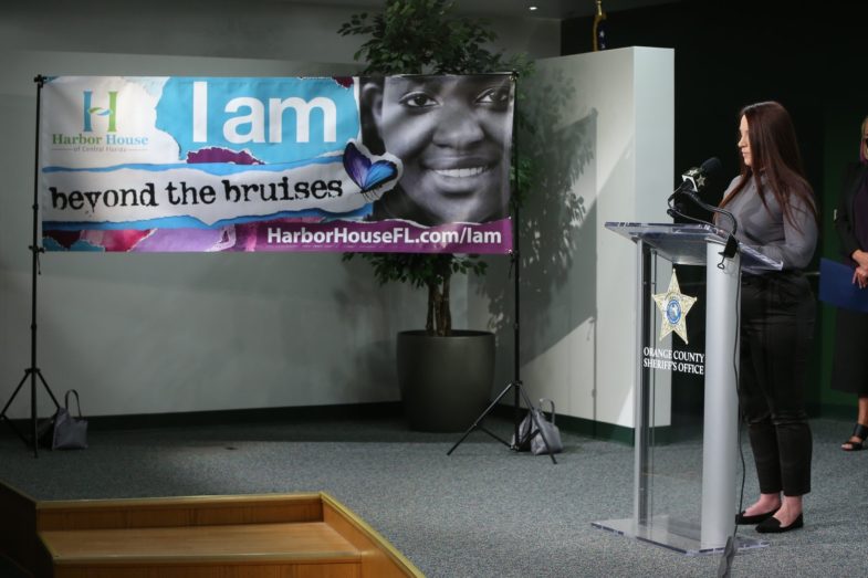 Woman speaks at podium in from of Domestic Violence Awareness Poster