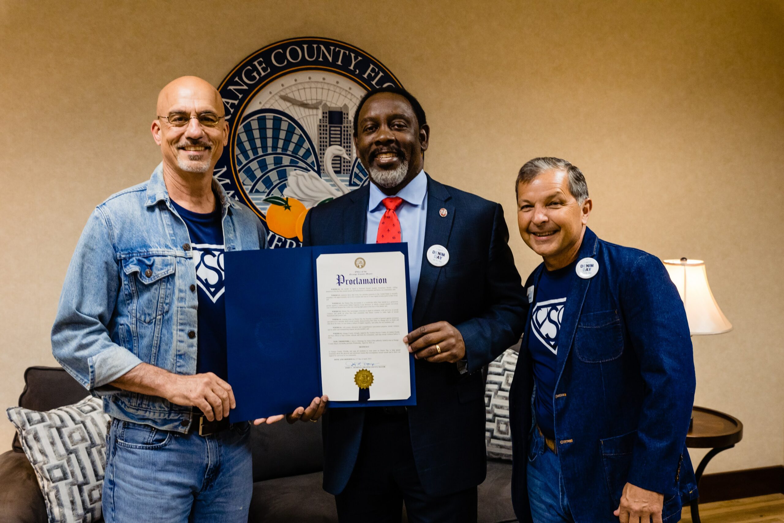 El Alcalde del Condado de Orange, Jerry Demings, sostiene una proclamación por el Denim Day 2023 acompañado por dos representantes del Centro de Servicios para Víctimas de la Florida Central.